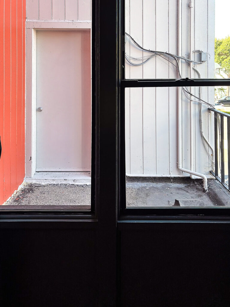 View from inside a La Luna Inn guest room window looking out onto a private gap between buildings, with red exterior wall and white siding visible outside