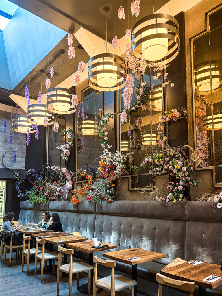 Interior of Blackwood Restaurant and Cafe in San Francisco's Marina District, featuring elaborate floral arrangements cascading from the ceiling and along the banquette wall, with drum pendant lighting and wood dining tables