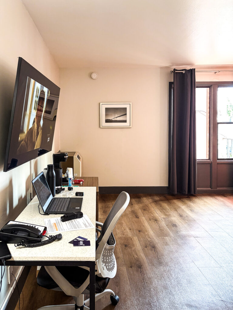 La Luna Inn guest room showing a work desk with laptop and hotel phone, wall-mounted TV, hardwood floors, dark curtains, window with natural light, and a black and white Golden Gate Bridge print on the wall
