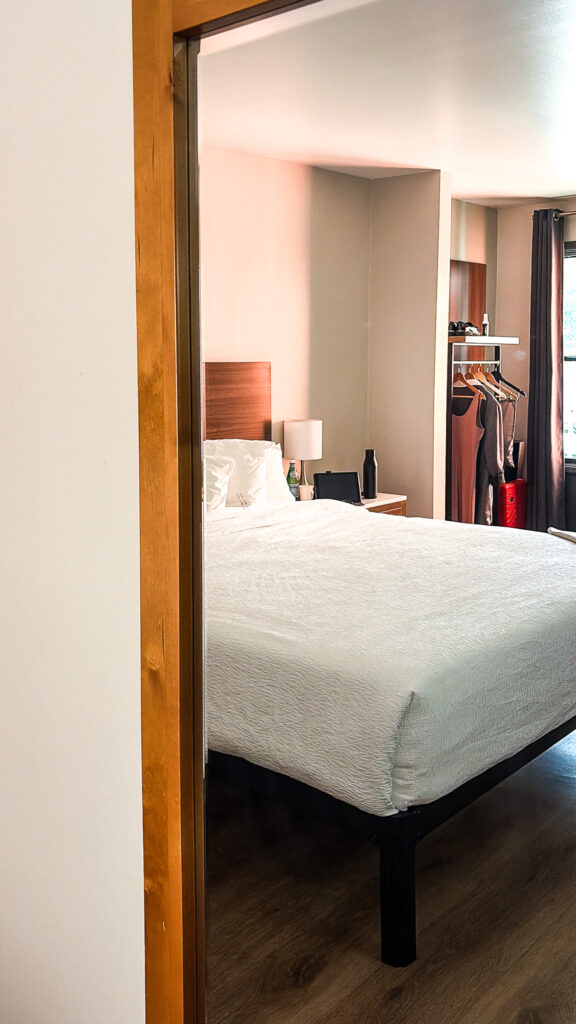 La Luna Inn guest room viewed through the doorway, showing a white bed with wood headboard, nightstand lamp, open clothing rack with hanging garments, hardwood floors, and dark curtains